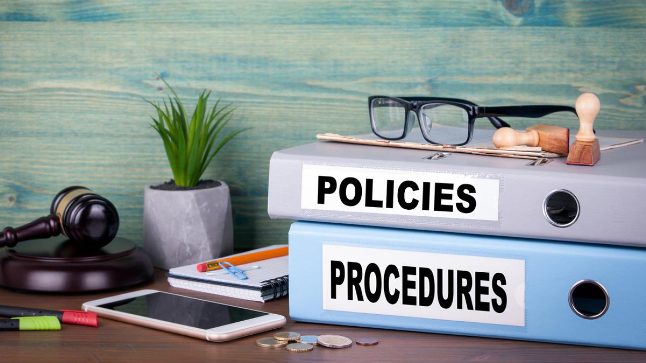 Image of a desk with items including two binders labeled &quot;POLICIES&quot; and &quot;PROCEDURES.&quot;